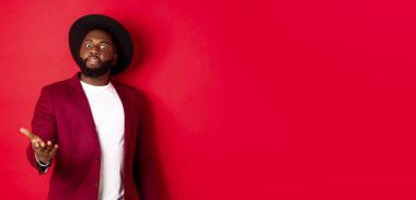 Şok olmuş Afro-Amerikan bir adam kameraya bakıyor ve üzgün bir şeye işaret ediyor, parti ceketi ve şapkası içinde duruyor, kırmızı arka plan.