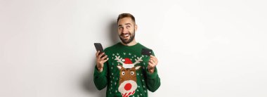 Yeni yıl, online alışveriş ve noel konsepti. Genç adam internetten hediye alıyor, elinde cep telefonu ve kredi kartı var, beyaz bir geçmişi var..