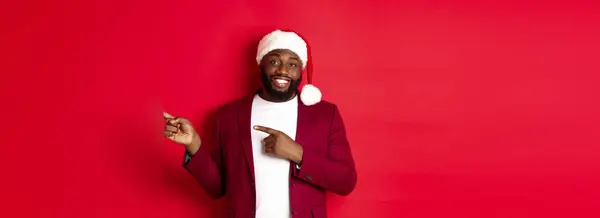 stock image Christmas, party and holidays concept. Smiling Black man with beard and santa hat, pointing fingers right at showing logo, standing over red background.