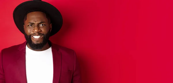 Stock image Close-up of Black man feeling uncomfortable, grimacing as watching something painful, cringe and frowning, standing over red background.