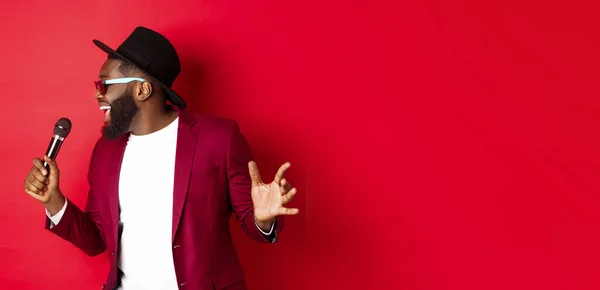 stock image Passionate black male singer performing against red background, singing into microphone, wearing party outfit, standing over red background.