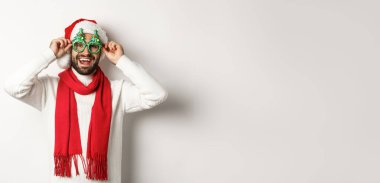 Noel, yeni yıl ve kutlama konsepti. Mutlu adam gülüyor, Noel Baba şapkası ve parti gözlüğü takıyor, beyaz arka planda duruyor..