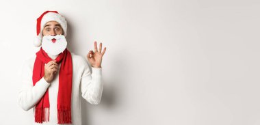 Noel partisi ve kutlama konsepti. Noel Baba şapkalı ve beyaz sakallı mutlu erkek manken, tamam jest gösteriyor, beyaz arka planda duruyor..