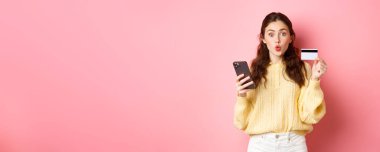 Teknoloji ve online alışveriş. Plastik kredi kartı gösteren, cep telefonu tutan, akıllı telefonla ödeme yapan, internetten sipariş veren, pembe arka planda duran heyecanlı kız..