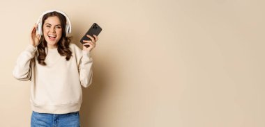 Happy caucasian woman listening music in headphones on smartphone app, dancing and having fun, standing over beige background.