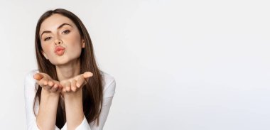 Feminine woman sending air kiss at camera, coquettish flirty pose, kissing, standing over white background. Copy space