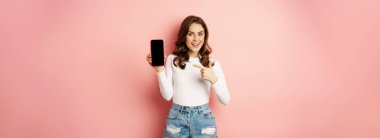 Stylish sexy woman showing mobile phone screen, smartphone app interface and smiling, demonstarting online shop, standing over pink background.