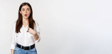 Portrait of young woman pointing at herself with disbelief, surprised face expression, being named or chosen, standing over white background.