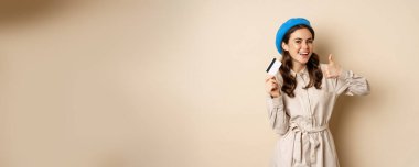 Concept of shopping and sale. Happy young woman in stylish clothes, showing credit card store discount and thumbs up, recommending, standing over beige background.