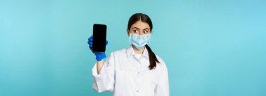 Young woman doctor in medical face mask and hospital uniform, showing mobile phone app, screen interface, online clinic concept, standing over torquoise background.