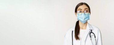 Medical worker, young doctor looking amazed and surprised, wearing face mask to prevent catching covid-19, standing over white background.