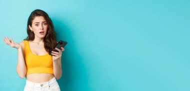 Teknoloji ve yaşam tarzı konsepti. Cazibeli kız akıllı telefon ekranını okuduktan sonra şaşkın görünüyor, kameraya şaşkın şaşkın bakıyor, mavi arka planda duruyor..