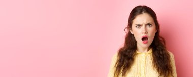 Şok olmuş ve incinmiş genç bir kadın, üzgün ve hayal kırıklığına uğramış bakıyor, soluk soluğa, yargılayıcı görünüyor, pembe arka planda duruyor..