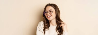 Close up portrait of stylish brunette woman in glasses, laughing and smiling, posing in eyewear against beige background.