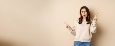 Confused girl shopper pointing sideways, making choice, decision between two variants, beige background. Copy space