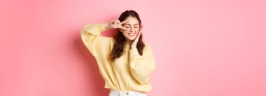 Güzel esmer kadın modelin görüntüsü, gülümseyen ve yüzüne dokunan, göze yakın V işaretini gösteren, mutlu bir şekilde gülen, pembe arka planda duran.