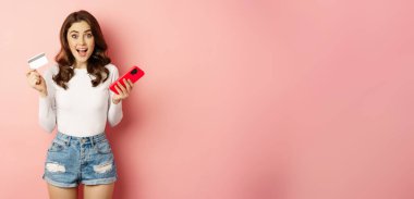 Enthusiastic brunette girl showing credit card and using mobile phone to order or pay, online shopping app, standing over pink background.