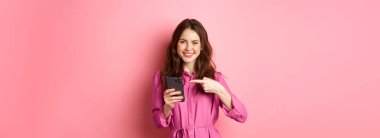 İnternette reklam yapan beyaz bir kız akıllı telefonuna işaret ediyor ve gülümsüyor. Tatmin olmuş, uygulama öneriyor, pembe arka planda duruyor..