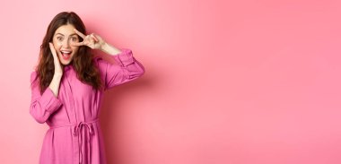 Pozitif esmer kız parti yapıyor, mutlu görünüyor, yüzüne dokunuyor ve V işareti gösteriyor, pembe arka plana karşı heyecanlı duruyor..