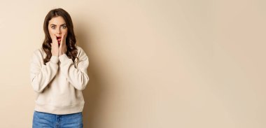 Portrait of shocked brunette woman drop jaw, gasping and staring speechless at camera, beige background. Copy space