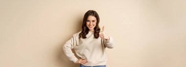 Beautiful young woman, student showing thumbs up in approval, recommending store, standing over beige background.