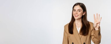 Portrait of corporate woman, saleswoman showing number four fingers and smiling, standing in suit over white background.