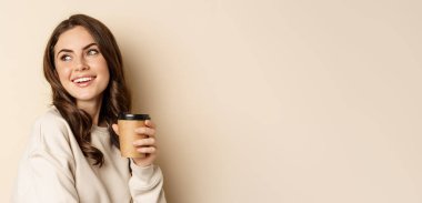 Takeaway and cafe concept. Beautiful feminine woman smiling, holding cup of coffee, posing against beige background. Copy space