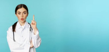 Young doctor, woman physician in white coat raising finger, pointing up, suggesting smth, has solution, revelation, standing over torquoise background.