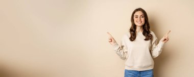 Cheerful caucasian woman showing directions, two ways, pointing sideways at variants, choices for customer, standing over beige background.
