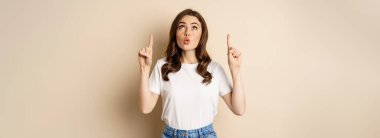 Surprised brunette girl showing promo, pointing and looking up with amazed face expression, standing over beige background.