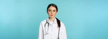 Medical staff and doctors concept. Young smiling female doctor, healthcare worker in white coat and stethoscope, looking confident, waiting patients, blue background.