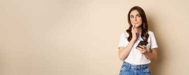 Girl thinks, holds mobile phone, looks up thoughtful, stands over beige background.