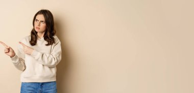 Woman pointing left and staring confused at banner copy space, strange logo, standing over beige background.