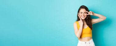 Temiz tenli ve mükemmel vücutlu güzel bir kadının portresi, yazlık kıyafetler giyiyor, barış işareti gösteriyor ve yüzlerine dokunuyor, mavi arka planda duruyor..