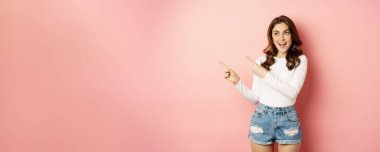 Cute smiling brunette girl, showing advertisement, pointing fingers left at banner, logo on empty pink space, studio background.