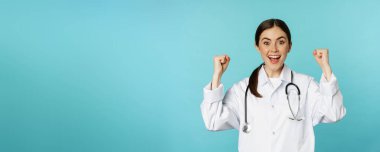 Enthusiastic doctor woman, rejoicing, winning and celebrating, achieve goal, standing in white coat against blue background. Copy space