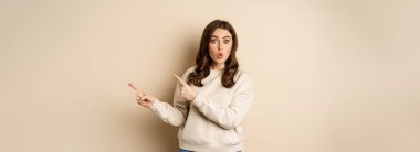 Surprised brunette woman pointing fingers right, showing advertisement and say wow impressed, standing over beige background.