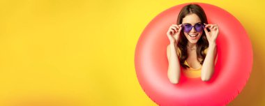 Close up portrait of stylish young woman, girl with swimming ring and summer sunglasses, ready for beach holiday, vacation, standing over yellow background.