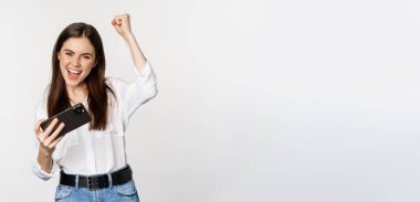 Happy brunette woman playing mobile video game, smiling and looking at screen excited, standing over white background.
