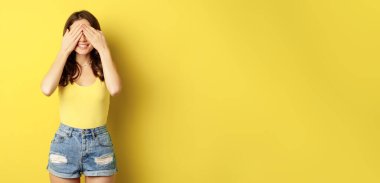 Portrait of brunette woman cover eyes with hands, blindfolded, smiling with anticipation, expecting smth, waiting for surprise, standing over yellow background.