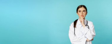 Young female doctor, hospital worker in white coat, thinking and looking away thoughtful, searching solution, standing over toquoise background.