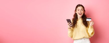 Teknoloji ve online alışveriş. Sipariş veren heyecanlı kız, kredi kartıyla ödeme yapan, cep telefonu tutan ve kameraya gülümseyen, pembe arka plan..