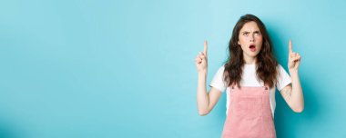 Şok olmuş genç bir kadın, somurtuyor, soluk soluğa, hayal kırıklığına uğramış ve gücenmiş bir şekilde bakıyor, saygısızca bir şey gösteriyor, mavi arka planda duruyor..