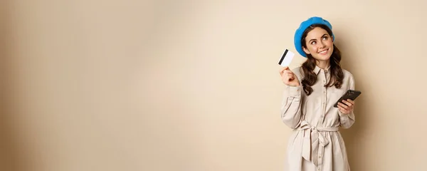 stock image Portrait of trendy woman in coat holding smartphone, showing credit card, shopping online, order something on mobile phone, standing over beige background.