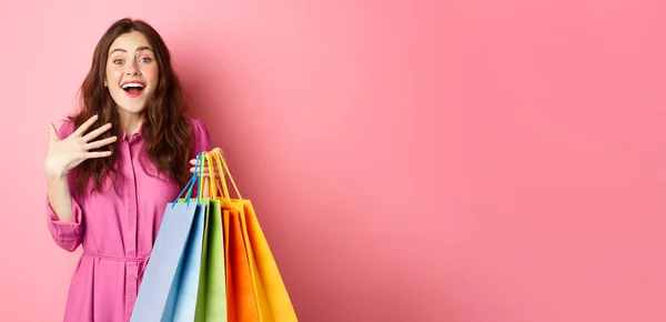 令人惊讶的年轻女子惊讶地喘息 手挽手 带着打折的微笑 提着购物袋 站在粉色的背景下 — 图库照片
