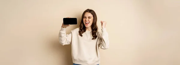 快乐的女孩 展示手机的水平屏幕和庆祝 欢呼与欢乐的脸 站在米色背景之上 — 图库照片