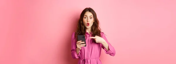 年轻的女人指着她的手机喘气 谈论智能手机应用 在网上展示一些有趣的东西 站在粉红的背景上 复制空间 — 图库照片