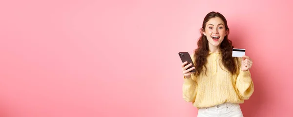 技术和网上购物 兴奋的女孩下订单 用塑料信用卡网上支付 拿着手机 对着相机微笑 粉红的背景 — 图库照片