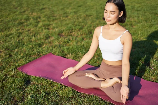 stock image Portrait of smiling asian woman meditating, doing yoga on fresh air, relaxing on rubber mat, exercising in park, breathing air, being calm.