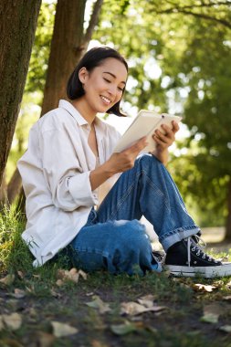 Güzel, genç Asyalı bir kız, öğrenci ağacın altında oturuyor ve kitap okuyor, gülümsüyor, dışarıda sıcak bir yaz gününün tadını çıkarıyor..